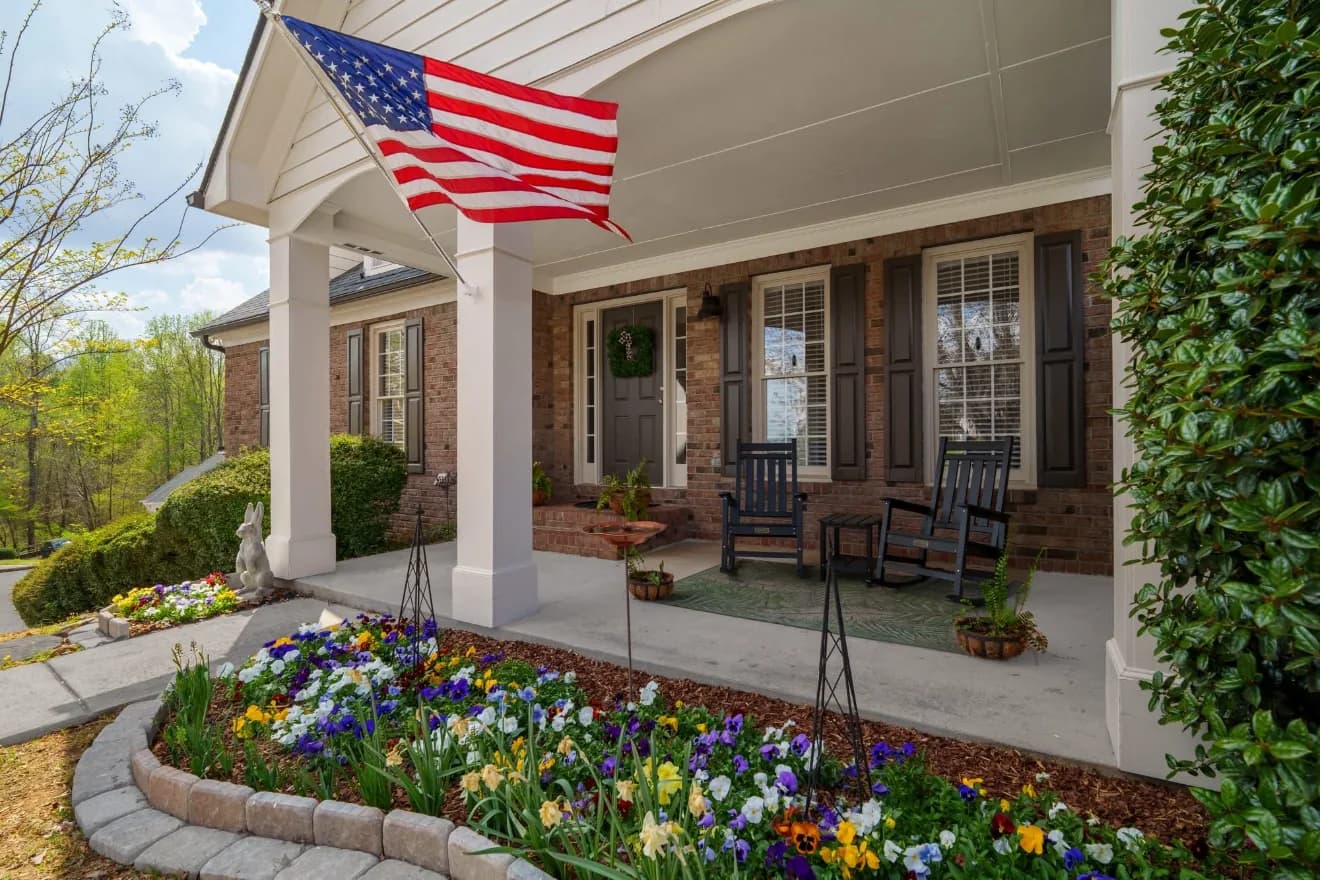 Covered front porch with rocking chairs, an American flag, and spring flower beds